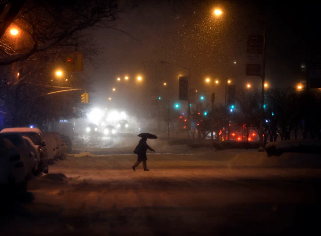 A silhouetted figure crosses Park Avenue in a snow storm with twinkling lights all around it and an umbrella.